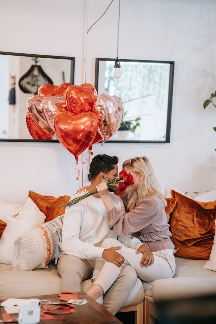 Interracial couple embracing with red roses and heart balloons on a cozy sofa, perfect for Valentine's Day.